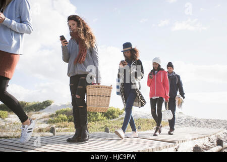 Fila di giovani amici adulti passeggiando lungo beach boardwalk lettura smartphone, Western Cape, Sud Africa Foto Stock