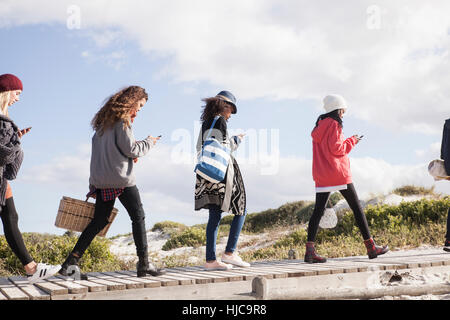 Fila di giovani amici adulti passeggiando lungo beach boardwalk lettura smartphone, Western Cape, Sud Africa Foto Stock