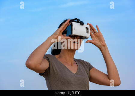 Donna (in 40s) indossando una realtà virtuale auricolare (VR) di maschera con il cielo a poche nuvole dietro di lei. Foto Stock