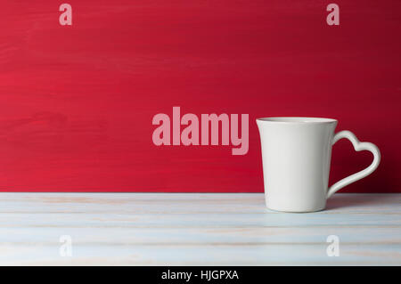 Tazza bianca con cuore maniglia nella parte anteriore rosso sfondo grunge sul tavolo bianco. Concetto di San Valentino. Foto Stock