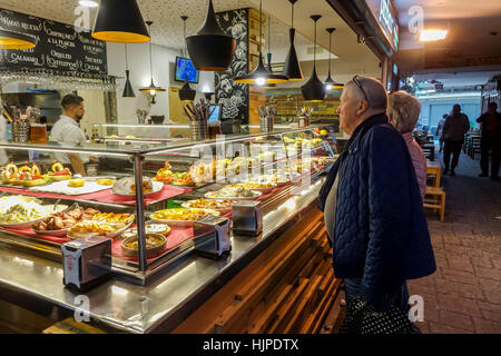 Tapas bar interno a Benidorm, Spagna. La vetrina in vetro mostra una varietà di pinchos, frutti di mare e crocchette. Selezione delle viste delle coppie di anziani al banco Foto Stock