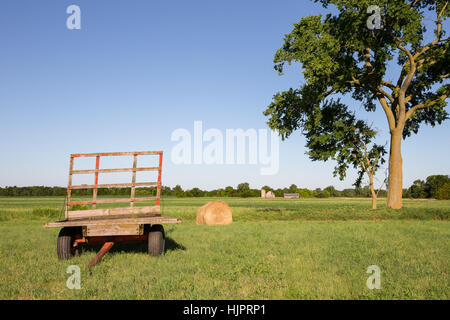 Carro autocaricante e balle di fieno in una fattoria campo sotto un grande albero. Copia dello spazio nel cielo se necessario e una fattoria nel lontano sullo sfondo. Foto Stock