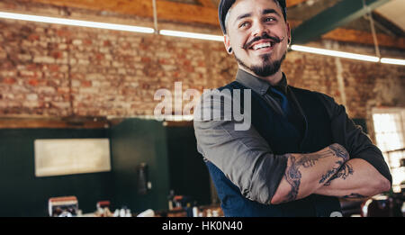 Close up di felice giovane maschio barbiere con cappuccio che guarda lontano e sorridente. Bello il parrucchiere in salone. Foto Stock