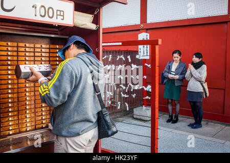 A cassetti di sinistra in un tempio contenente Omikuji fortune raccontando le carte. Ragazzo cerca il suo Omikuji. Sullo sfondo le ragazze leggere loro Omikuji. Di Senso-ji T Foto Stock