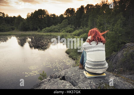 Ragazza seduta sulle pietre costiere vicino lago ancora nel tramonto, vista posteriore Foto Stock