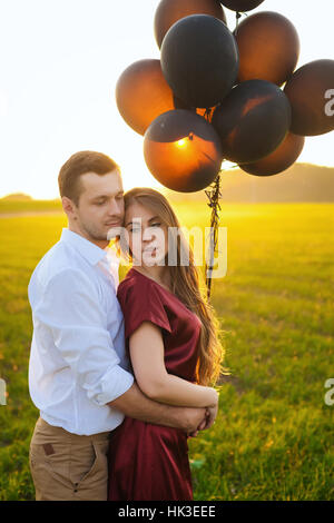 Ragazzo in camicia bianca e una ragazza in abito rosso avvolgente al tramonto. Nelle loro mani sono nero palloncini. Sono felice in lovers day Foto Stock