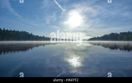 Brillanti e luminose a metà estate alba sulle sponde di un lago. Acqua calda e lo scambiatore di calore aria allo spuntar del giorno crea densa nebbia patch. Ancora acqua su acque calme. Foto Stock