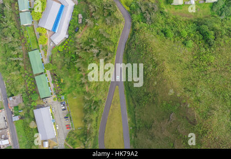 Una vista aerea dall'alto verso il basso mostra una stretta e tortuosa strada asfaltata che attraversa un lussureggiante paesaggio verde. Sul lato sinistro della strada, ci sono diversi edifici con tetti grigi e verdi, tra cui Foto Stock