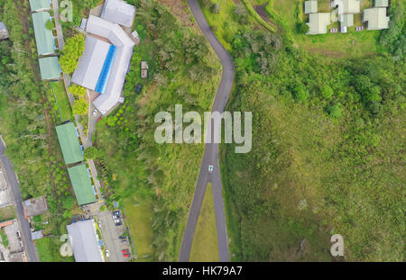 Una vista aerea dall'alto verso il basso mostra una stretta e tortuosa strada asfaltata che attraversa un lussureggiante paesaggio verde. Sul lato sinistro della strada ci sono diversi edifici con tetti verdi e grigi, alcuni con Foto Stock