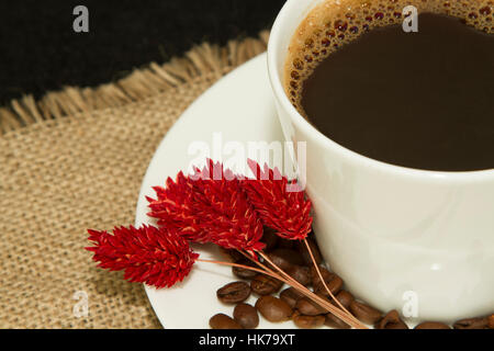 White tazza di caffè con i chicchi di caffè sul piattino e asciugare fiore rosso sul panno di lino Foto Stock