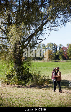 Una giovane donna e uomo in piedi accanto a un albero in un campo che si baciano. Foto Stock