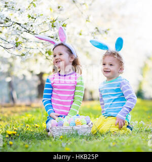 I bambini su Easter egg hunt nel giardino fiorito. I bambini con orecchie di coniglietto alla ricerca di uova colorate in caduta di neve prato di fiori Foto Stock