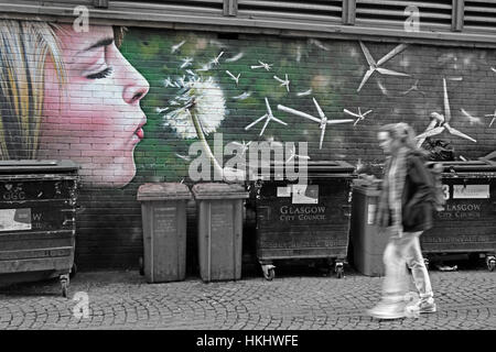 Graffiti a Glasgow dietro le quinte, una donna bionda soffia un tarassolo, nelle turbine eoliche, generando energia verde, vicino ai bidoni del grano Foto Stock