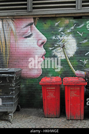 Donna bionda che soffia i semi da un orologio di dente di leone, sopra i bidoni dei rifiuti, Glasgow, Scozia, UK Foto Stock