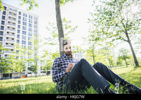 Giovane uomo seduto in un parco e la scrittura nel blocco note, Monaco di Baviera, Germania Foto Stock