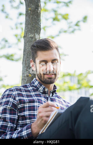 Giovane uomo seduto in un parco e la scrittura nel blocco note, Monaco di Baviera, Germania Foto Stock