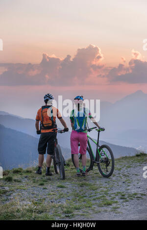 Vista posteriore della giovane coppia di mountain bikers in piedi nel paesaggio alpino e cercando di visualizzare durante il tramonto, Zillertal, Tirolo, Austria Foto Stock