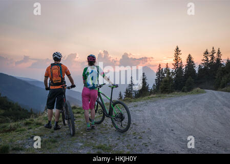 Vista posteriore della giovane coppia di mountain bikers in piedi nel paesaggio alpino e cercando di visualizzare durante il tramonto, Zillertal, Tirolo, Austria Foto Stock