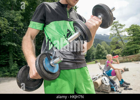 Giovane uomo il sollevamento pesi mentre la donna è acqua potabile in background, Kampenwand, Baviera, Germania Foto Stock