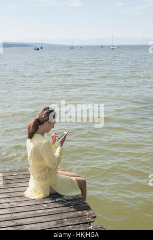 Donna che utilizza smart phone e seduto sul molo al lago Ammersee, Alta Baviera, Germania Foto Stock