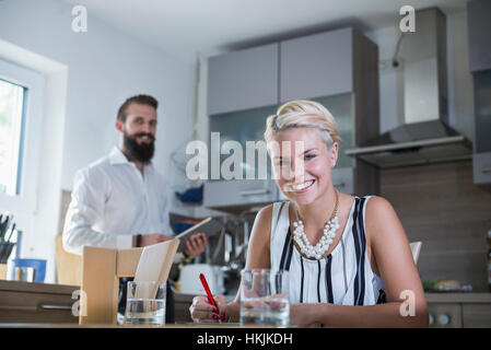 Giovane donna iscritto al tavolo di cucina e uomo in background con tavoletta digitale, Baviera, Germania Foto Stock