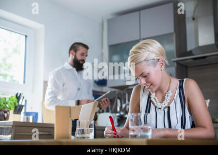 Giovane donna iscritto al tavolo di cucina e uomo in background con tavoletta digitale, Baviera, Germania Foto Stock