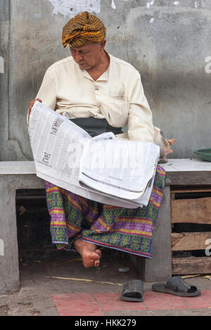 Uomo seduto su una panchina di pietra leggendo un giornale a Denpasar, Bali, Indonesia Foto Stock