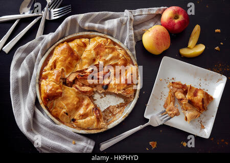 Un rustico in casa torta di mele con un serving rimosso. Forche, un tovagliolo e le mele sono dispersi in preparazione. Foto Stock