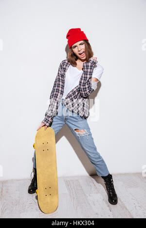 Foto di emotivo giovane signora vestita in camicia in una gabbia di stampa cappello da indossare in piedi isolato su sfondo bianco azienda skateboard Foto Stock