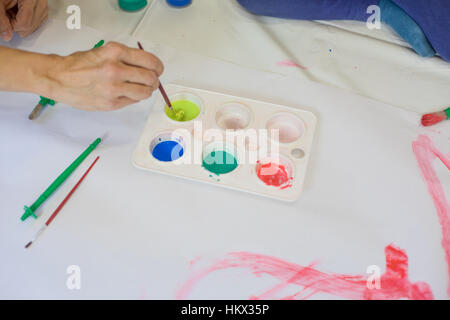 Donna mano con pennello Acquerelli preparazione giallo, blu, verde e rosso seduta su bianco in plastica e carta accanto al bambino per la verniciatura Foto Stock