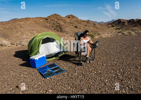 Donna nel deserto (montagna) che gioca con il suo smartphone mentre si ricarica con un pannello solare portatile e pieghevole. Foto Stock