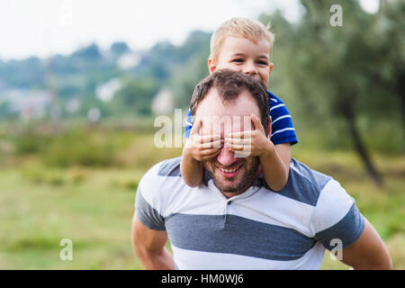 Il ragazzo si chiude gli occhi mani padre. Happy little boy godendo con riding sul padre indietro. Happy Family Portrait. Ridere papà con little boy godendo di nat Foto Stock