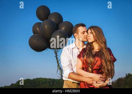 Uomo in camicia bianca e la donna in abito rosso che abbraccia oltre il cielo blu. Nelle loro mani sono nero palloncini. Sono felice nel giorno di San Valentino Foto Stock