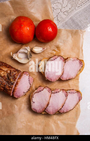 Fette affumicato con spezie e erbe aromatiche a base di carne o prosciutto su marrone carta da imballo. Pomodori, aglio e piccoli spuntini di pane e carne e il vecchio coltello vintage. Foto Stock