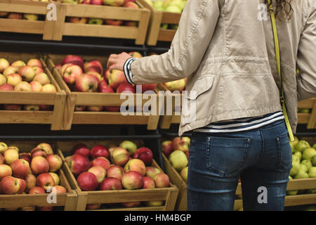 Ragazza in giacca beige e blu jeans raccoglie le mele al supermercato. Ragazza visto dal retro. Sullo sfondo è possibile vedere le scatole di mele. Foto Stock