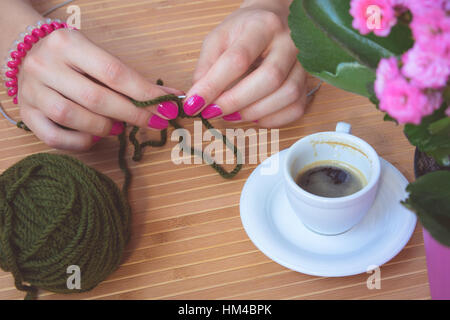 Le mani delle donne con viola manicure vengono lavorati a maglia razze metalliche di un tavolo di legno. Sul tavolo è una tazza di caffè, Vaso di fiori e una palla verde di lana ya Foto Stock