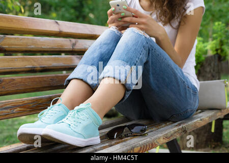 Ragazza in jeans si siede su una panchina nel parco e utilizzando un telefono cellulare Foto Stock