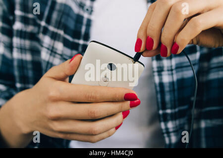 Donna in plaid shirt e manicure rosso consentono di collegare le cuffie al bianco telefono cellulare close-up Foto Stock