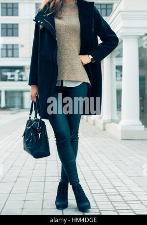 Dettagli di abbigliamento femminile. Una donna in un maglione, manto nero e pantaloni tenendo un borsette passeggiando per la città. Foto Stock