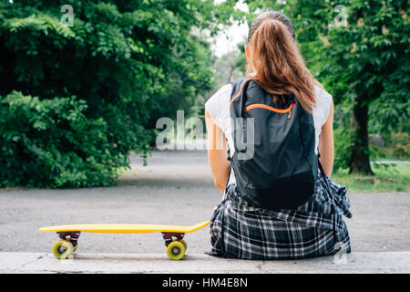 Vista dal retro della ragazza in jeans, t-shirt, sneakers e uno zaino, che siede sui gradini accanto al suo skateboard giallo nel parco Foto Stock