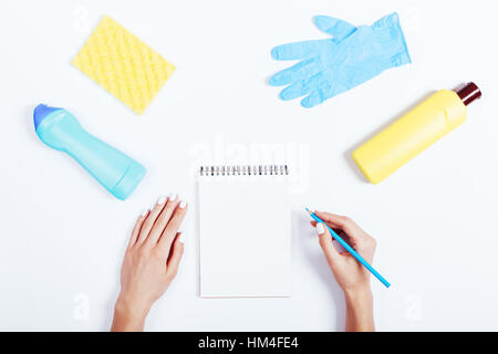 Mani femminili la scrittura nel blocco note, giallo e blu bottiglia di detersivo e spugna su sfondo bianco, vista dall'alto Foto Stock