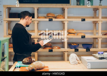 Vista laterale di baker iscritto sulla clipboard mentre in piedi da ripiani in forno Foto Stock