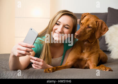 Ragazza adolescente (16-17) giacente sul letto con brown Vizsla cane e tenendo selfie Foto Stock