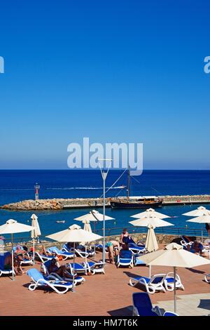 I turisti la rilassante Hotel Mirabello terrazza per prendere il sole con il porto verso la parte posteriore, Agios Nikolaos, Creta, Grecia, l'Europa. Foto Stock