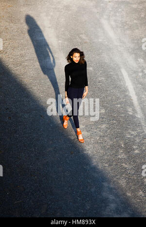 Ritratto di fiducioso giovane donna adulta vestiti di nero e arancione scarpe camminando durante la colata lunga ombra, ad alto angolo di visione Foto Stock