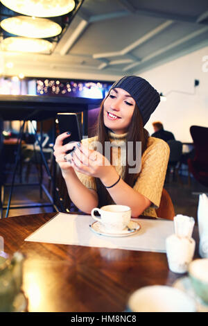 Ritratto di bella ragazza in hat usando il suo telefono cellulare in cafe. Tonned. Messa a fuoco selettiva. Foto Stock