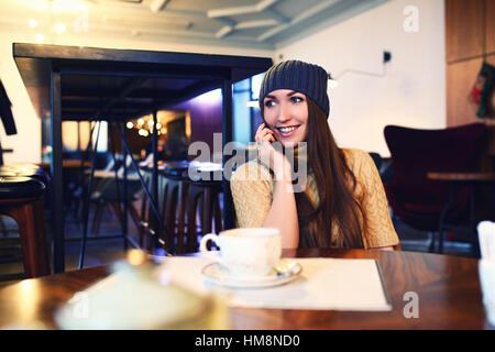 Ritratto di bella ragazza in hat usando il suo telefono cellulare in cafe. Tonned. Messa a fuoco selettiva. Foto Stock