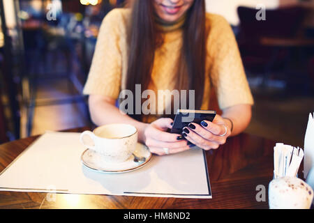 La donna la digitazione di un messaggio di testo sul telefono intelligente in un cafe'. Immagine ritagliata della giovane donna seduta al tavolo di un caffè utilizzando il telefono cellulare. Tonned. Messa a fuoco selettiva. Foto Stock