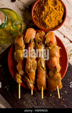 Primo piano di alcune materie Pollo speziato spiedini di carne in un piatto di terracotta, su una tavola in legno rustico, accanto a una ampolla di vetro con olio di oliva e una ciotola wit Foto Stock