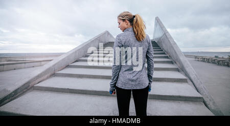 Vista posteriore del colpo di giovane donna in piedi da una scale prima di allenamento. Fitness modello femminile in piedi da passi in mattina. Foto Stock